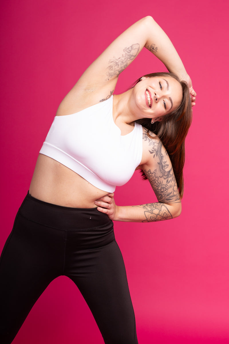 A smiling woman in a white sports bra and black leggings poses in a side stretch against a solid bright pink background.